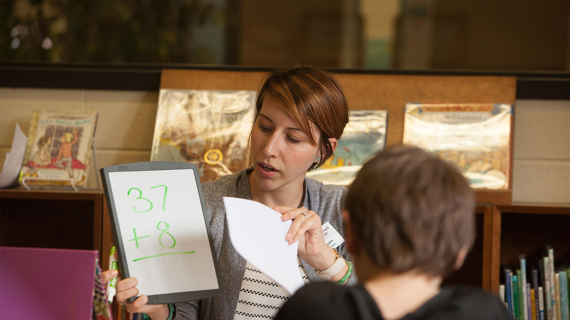 An elementary school teacher holds up a markerboard with a math problem for her students to solve.