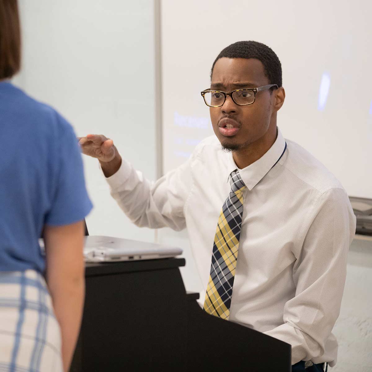 A teacher wearing a white, collared shirt with a tie speaks to a student during class.
