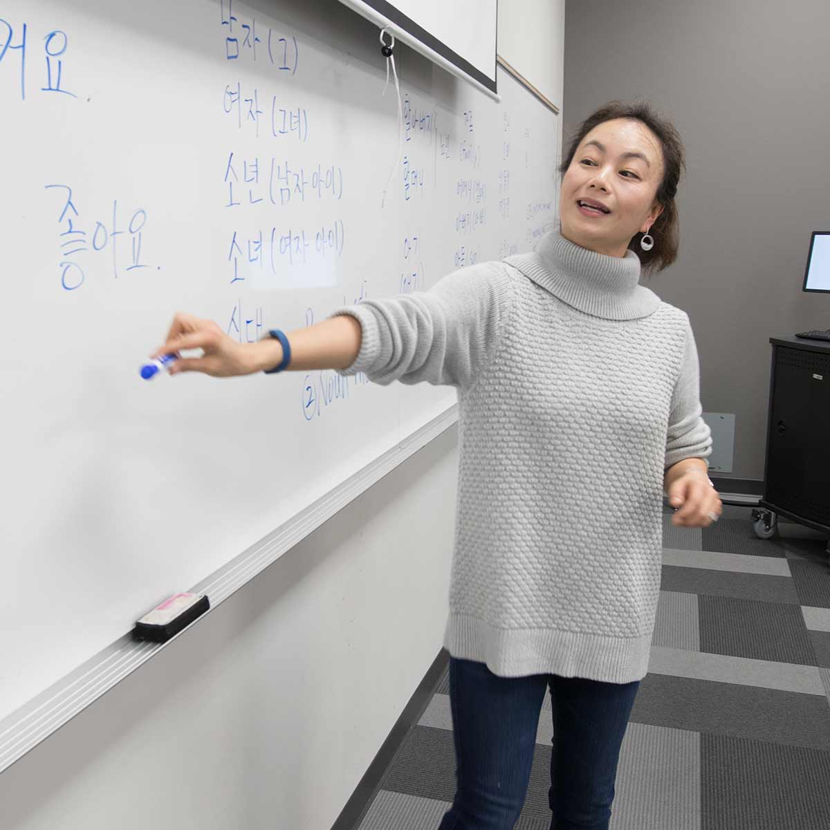 A language teacher holds up a blue marker to point to Chinese words and letters on a dry erase board during class.