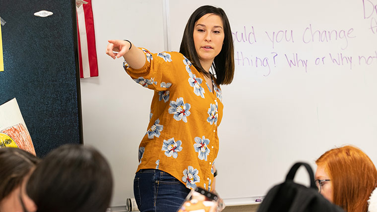 A teacher points to something while giving instructions at the front of the classroom.