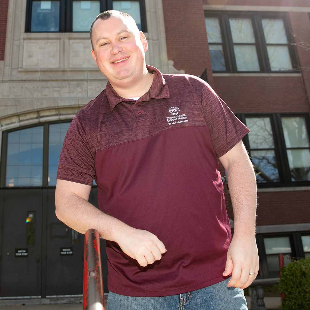A teacher wearing a maroon Missouri State polo stands outside in front of their school's main entrance.
