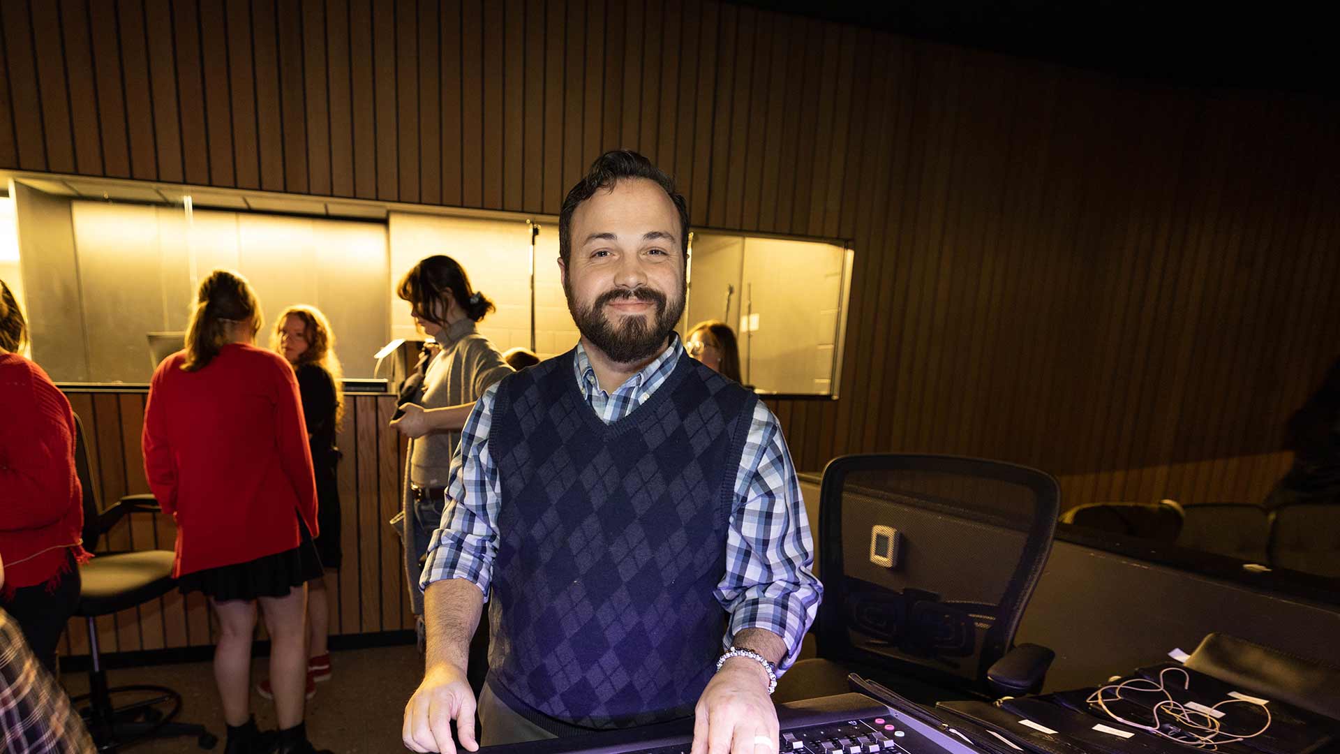 A theater teacher smiles from behind a soundboard during class. Students are seen behind him.