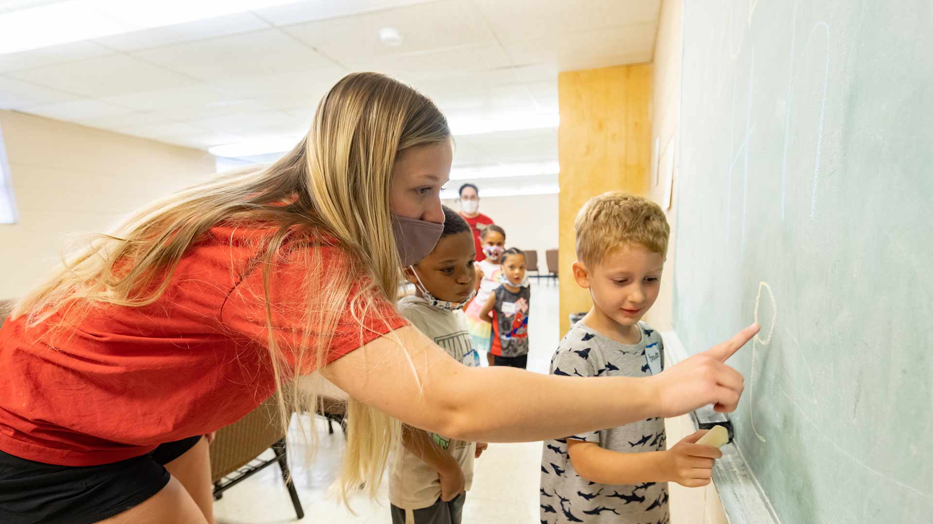 A teacher helping an elementary student write on a chalkboard in a classroom.