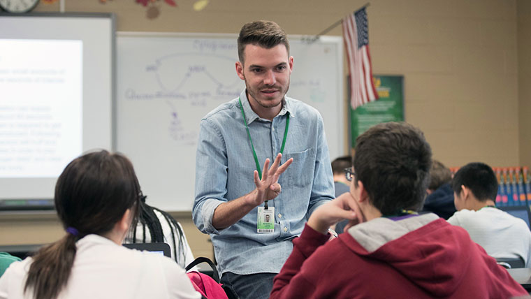 A teacher within a classroom holding up four fingers while explaining a lesson to two middle school students