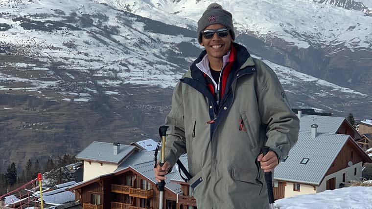 An education broad student on a ski slope in France. He's holding his set of skis and there are snow-covered mountains in the background.