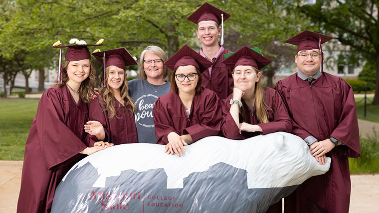 A group of Missouri State education graduates, dressed in their gradation regalia, gather around the MSU College of Education's Bear statue for a group photo.