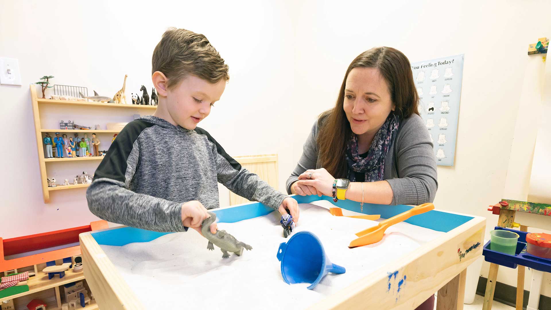 Toddler playing in an indoor sandbox with a dinosaur and motorcycle as a teacher observes. 
