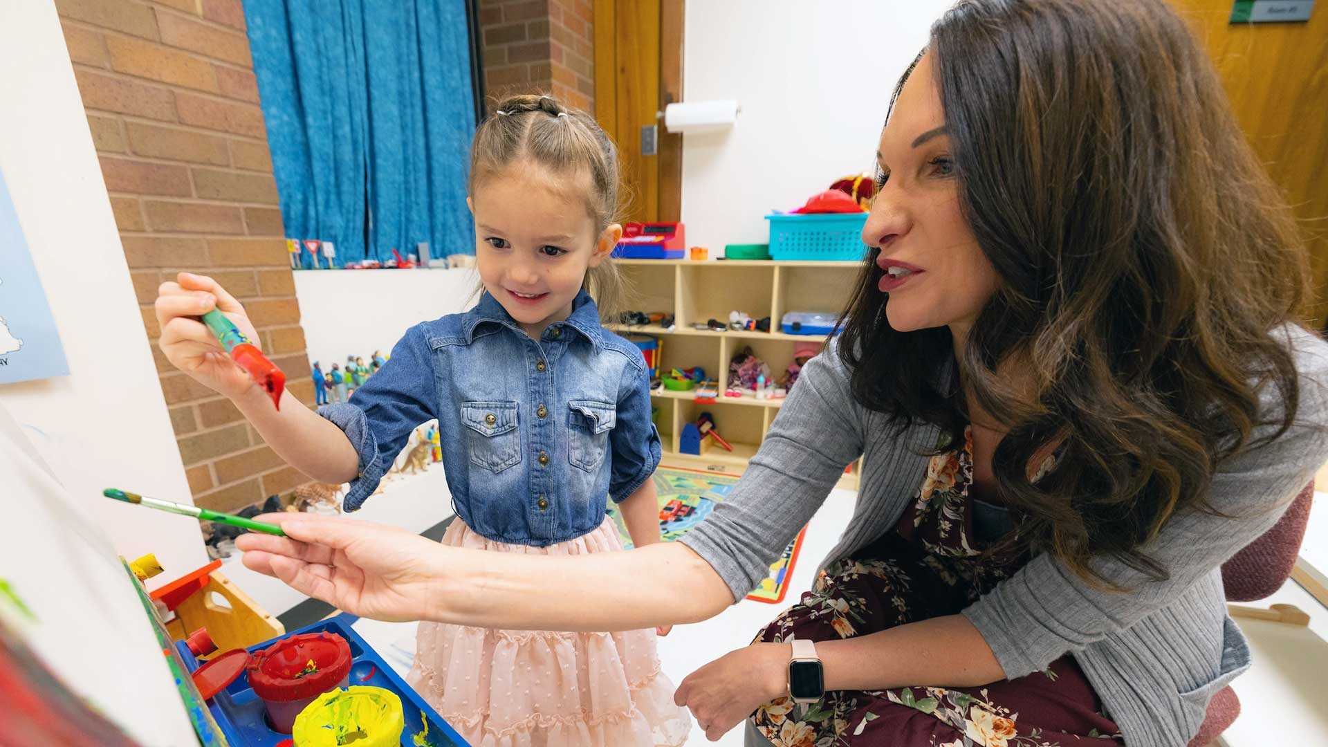 Teacher painting at an easel with a young student. 